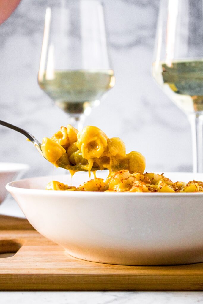 Head on view of a white bowl with a fork pulling Daiya mac and cheese recipe out of it with a glass of wine in the background