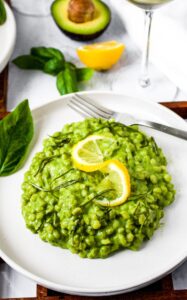 Overhead photo of a rount white plate topped with creamy avocado pesto risotto topped with a twisted lemon wedge