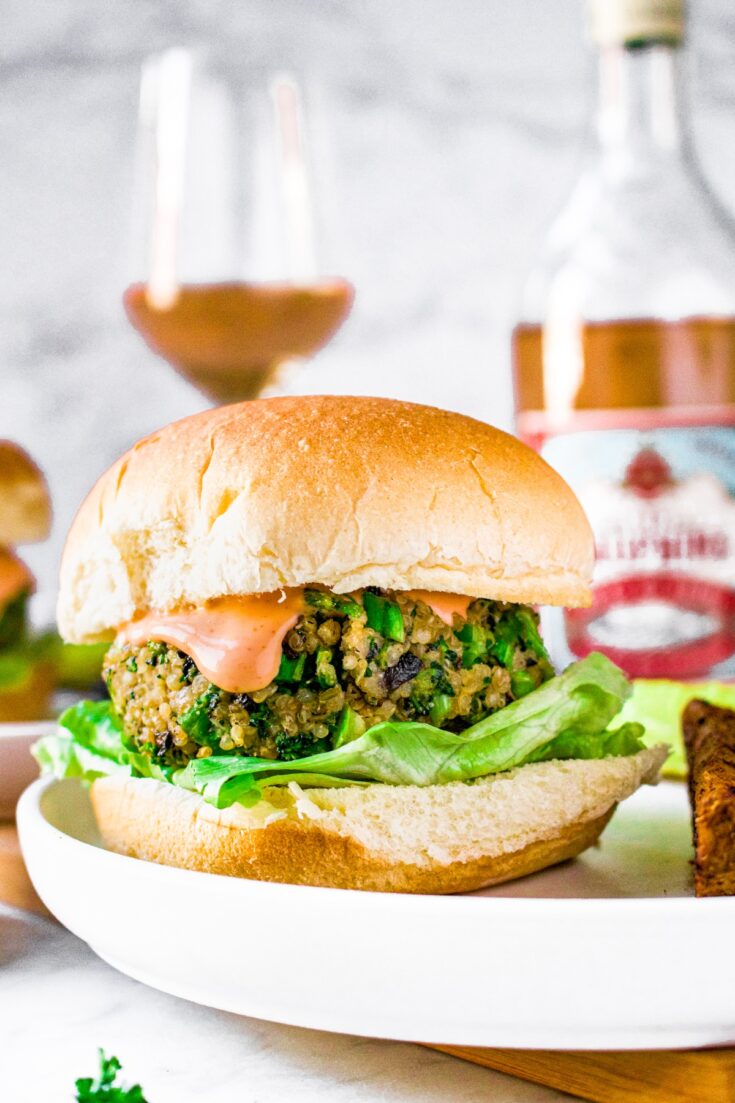 Head on shot of a mushroom quinoa broccoli burger on a bun with sriracha aioli dripping down the side. A bottle and glass of French rose is in the background.