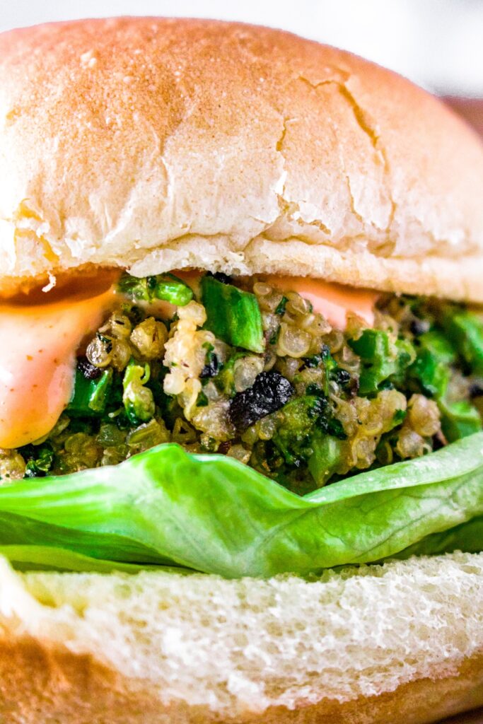 Close up head on shot of a broccoli quinoa burger on a bun with a crisp leaf of lettuce and creamy sriracha sauce dripping down the side.