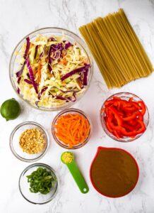 An overhead shot of the ingredients you need for cold peanut lime noodles: noodles, coleslaw mix, shredded carrots, sliced red peppers, scallions, ginger, peanut sauce or satay sauce, a lime, and chopped peanuts.