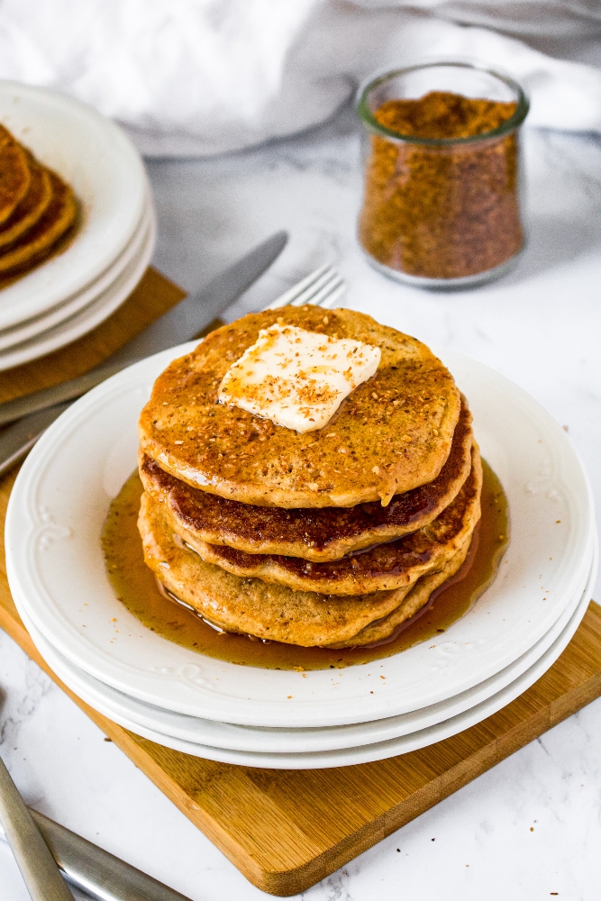 A stack of vegan pancakes with coconut bacon stacked on a small white plates topped with a square of vegan butter and a drizzle of maple syrup.