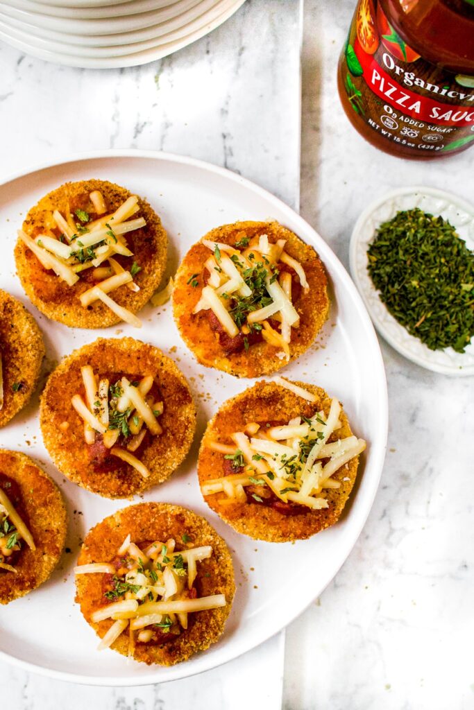 Overhead shot of vegan polenta pizza bites appetizers on a plate - breaded polenta circles topped with pizza sauce, melted vegan cheese, and Italian herbs.
