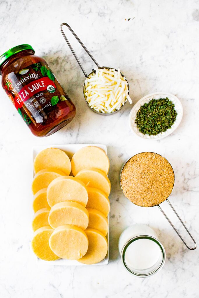 An overhead photo showing the ingredients you need to make vegan polenta pizza bites: a tube of polenta cut into circles, non dairy milk, bread crumbs, vegan mozzarella, dried herbs, and pizza sauce