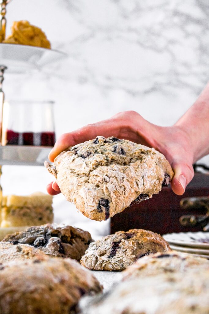 Head-on shot of a hand reaching in to pick up a dairy free blueberry scone. There is a 3-tiered tea tray to the left and more scones surrounding everything.