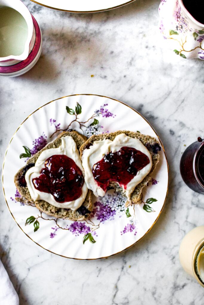 Overhead shot of a small china plate with delicate purple flowers painted on it topped with a bluberry scone sliced in half and topped with vegan clotted cream and strawberry jam.