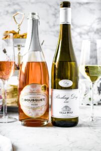 Head-on shot of two bottles of one - one is a sparkling rose from Domaine Bousquet. The other is a dry riesling from Villa Wolf. There is a glass next to each bottle filled with the wine. In the background, it's set up for an afternoon tea with a 3-tier tray and tea pot.