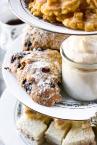 Overhead shot of a 3-tier tea tray with finger sandwiches on the bottom, blueberry scones and vegan clotted cream in the middle, and dairy free cheese straws on top. The focus is on the scones and clotted cream. There is a cup of hot tea in the background.