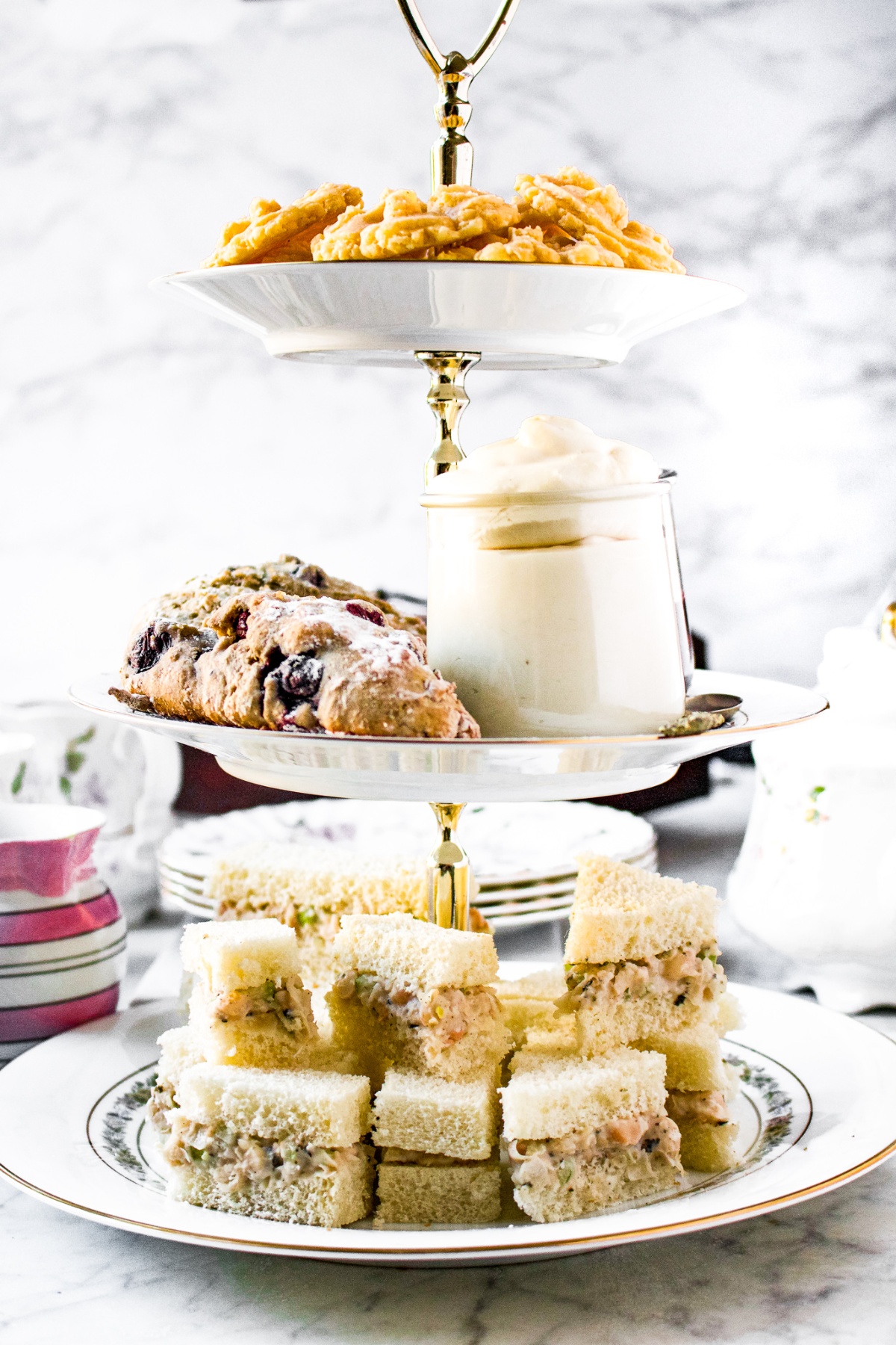 Head on shot of a 3-tier tea tray with finger sandwiches on the bottom, blueberry scones and vegan clotted cream in the middle, and dairy free cheese straws on top. There is a pot of tea and non dairy creamer in a pink and white striped pitcher in the background set up for an afternoon tea party.
