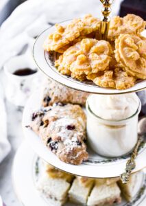 Overhead photo of a 3-tier tea tray with finger sandwiches on the bottom, blueberry scones and vegan clotted cream in the middle, and dairy free cheese straws on top. There is a cup of black tea in the background.