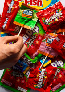 Overhead shot of a pile of vegan candy like Fun Dip, Skittles, and Jolly Rancher Lollipops. A hand is reaching in to grab one of the lollipops.