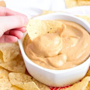 Overhead close up view of a chip being dipped into a creamy vegan queso sauce in a round white bowl