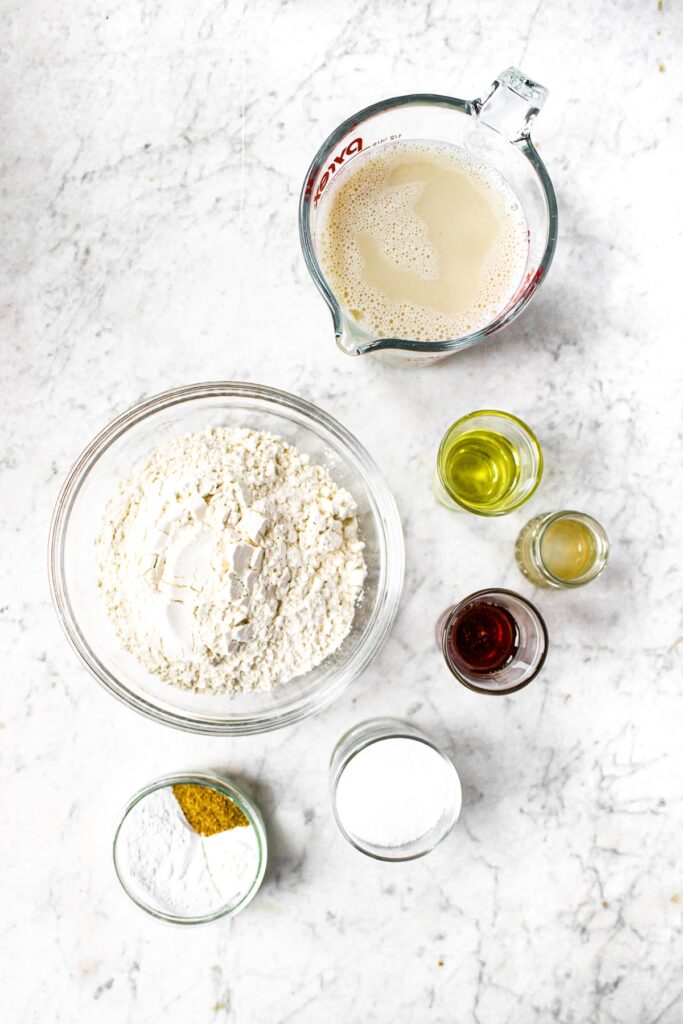 An overhead photo of all the ingredients you need to make plant-based waffles: flour, non-dairy milk, apple cider vinegar, ground flaxseed, baking powder, salt, sugar, oil, maple syrup, nutmeg and cinnamon