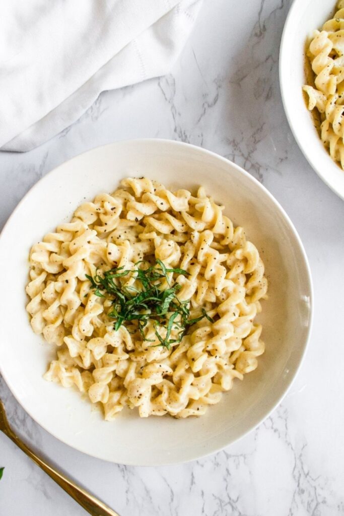Overhead view of a round white bowl filled with creamy vegan pasta in garlic sauce topped with fresh basil and ground black pepper