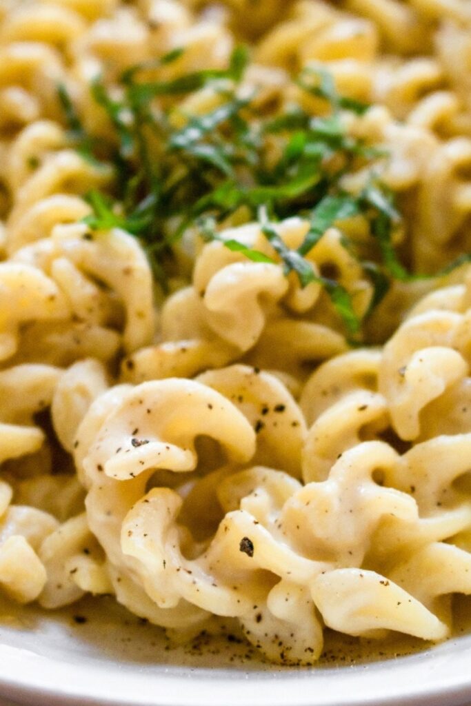 Overhead close up view of rotini pasta in a white bowl covered in creamy vegan garlic sauce and sprinkled with black pepper