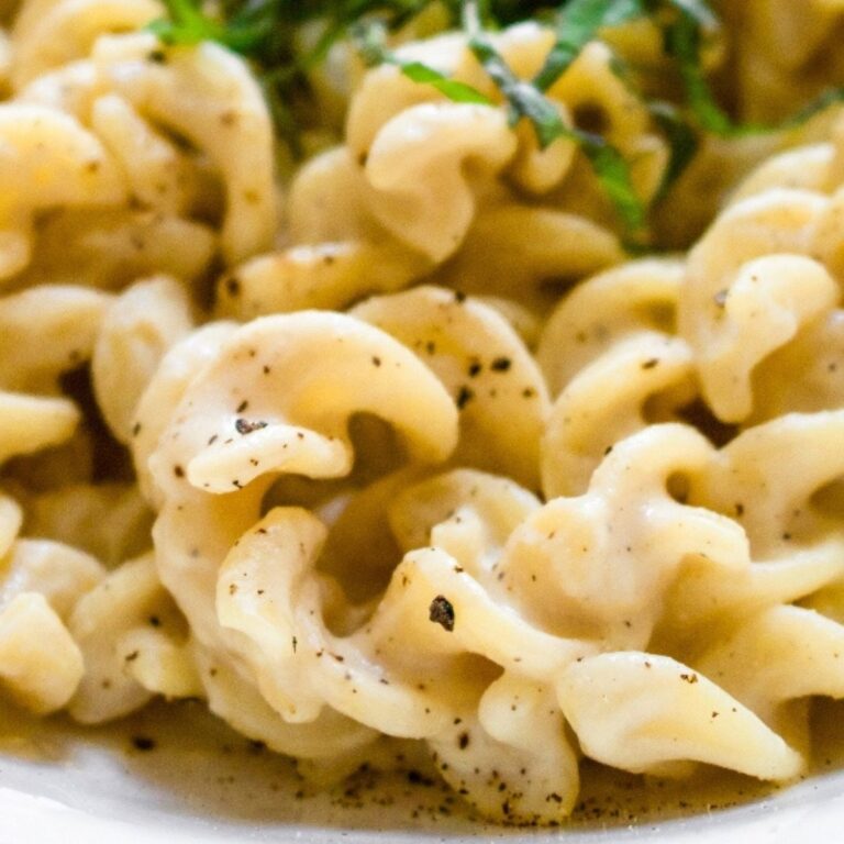 Overhead close up view of rotini pasta in a white bowl covered in creamy vegan garlic sauce and sprinkled with black pepper
