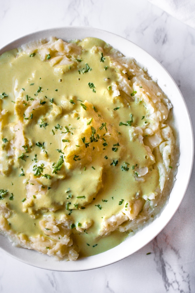 Overhead photo of a round white bowl of mashed potatoes drenched in vegan gravy and sprinkled with fresh minced herbs