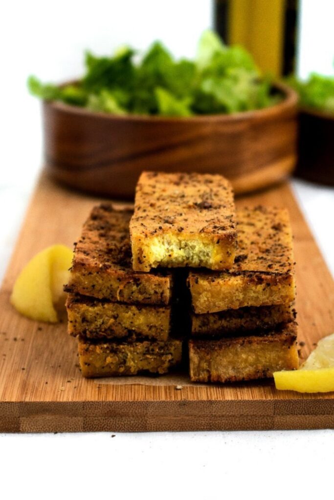 Head on view of a stack of seven baked lemon pepper tofu cutlets with a bite taken out of the top one