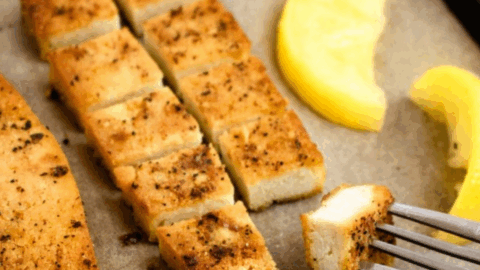 Overhead view of a lemon pepper tofu cutlet on a parchment lined baking sheet cut into small squares with a fork going into the square closest to the camera