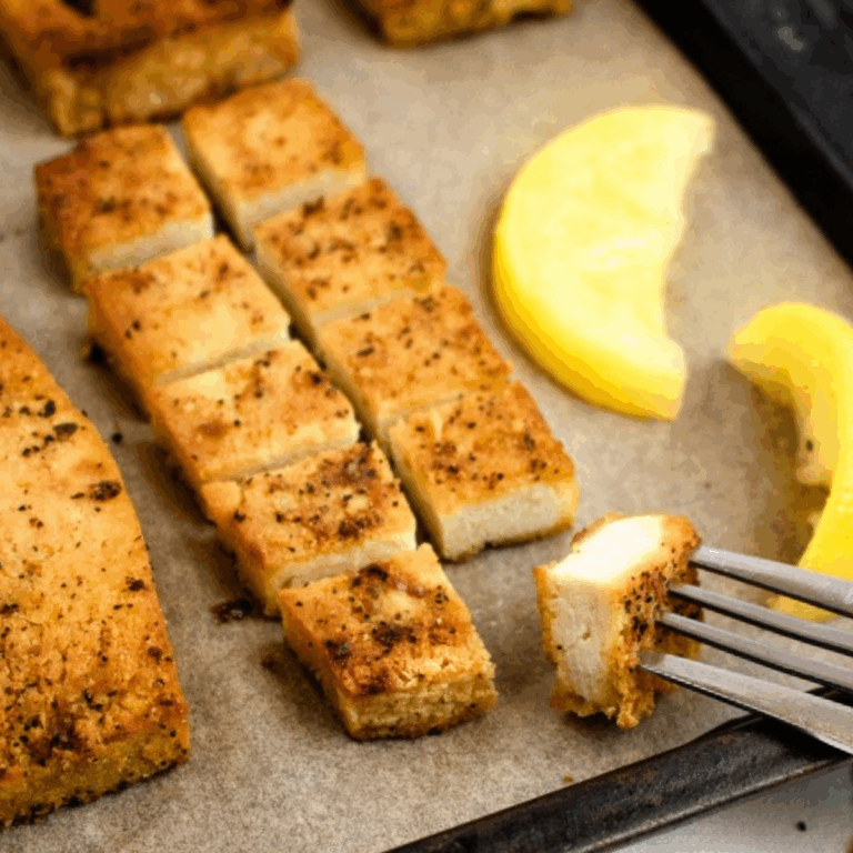 Overhead view of a lemon pepper tofu cutlet on a parchment lined baking sheet cut into small squares with a fork going into the square closest to the camera