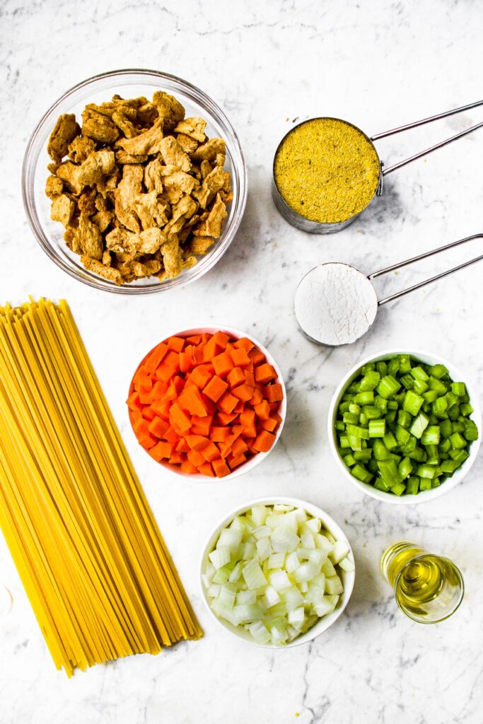 Overhead photo of the ingredients you need for vegan chicken noodle soup: spaghetti, diced carrots, celery, and onions, veggie chicken (soy curls), all purpose flour, no chicken bouillon powder, and olive oil
