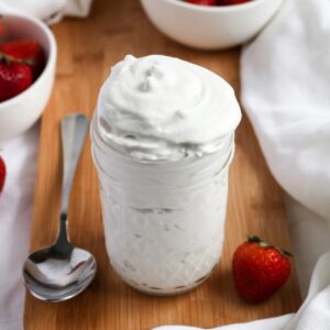 Overhead view of a clear glass jar filled with vegan whipped cream on a wood plank with a strawberry to the right and spoon to the left