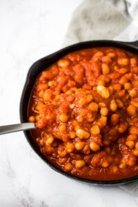 Vegan baked beans in a cast iron skillet with a spoon lifting some out. This delicious, healthy take on classic baked beans is filled with flavor but uses simple ingredients (and no ketchup!) on the stove.