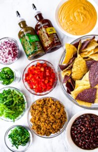 An overhead shot of the ingredients in dairy free and vegetarian loaded nachos - walnut meat, black beans, scallions, shredded lettuce, jalapenos, diced tomatoes and red onion, salsa verde and hot sauce, dairy free nacho cheese sauce, and chips.