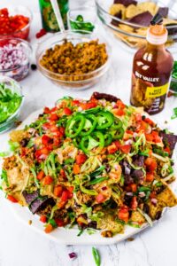 A photo of a big pile of vegan nachos on a white plate piled with spicy beef-style walnut meat, dairy free nacho cheese sauce, diced tomatoes and red onion, shredded lettuce, and sliced jalapenos. Extra toppings in clear bowls can be seen in the background.