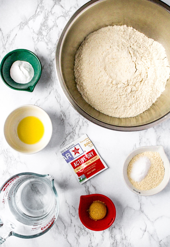 Overhead shot of the ingredients you need to make homemade bagels: all-purpose flour, baking soda, active dry yeast packet, warm water, olive oil, salt, and light brown sugar.