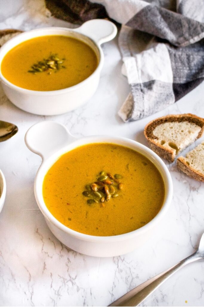Overhead view of two round white bowls filled with creamy vegan pumpkin soup topped with toasted pepitas next to a slice of pumpernickel bread