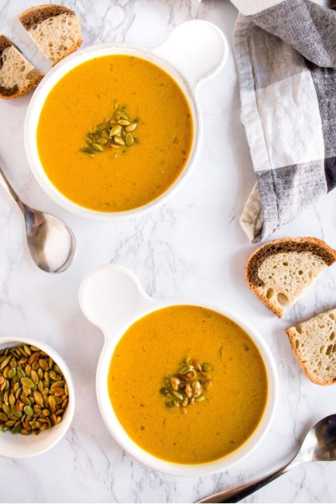 Overhead view of two round white bowls filled with spicy vegan pumpkin soup topped with toasted pumpkin seeds next to a slice of rye bread