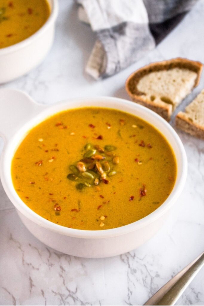 Overhead view of a round white bowl filled with vegan curried pumpkin soup topped with spicy red pepper flakes and toasted pepitas next to a slice of pumpernickel rye swirl bread