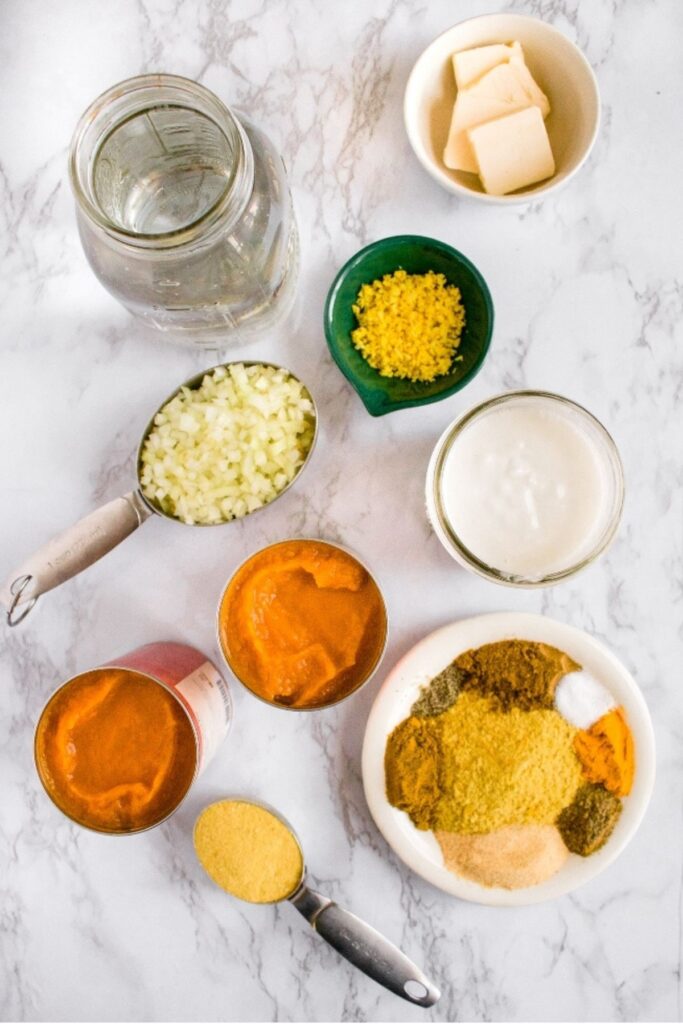 Overhead view of various soup ingredients in bowls and measuring cups sprawled out on a grey marble countertop such as cans of pumpkin puree, diced onion, minced garlic and ginger, spices on a round white plate, a clear jar of water, vegan cream in a glass, and dairy-free butter slices in a round white bowl