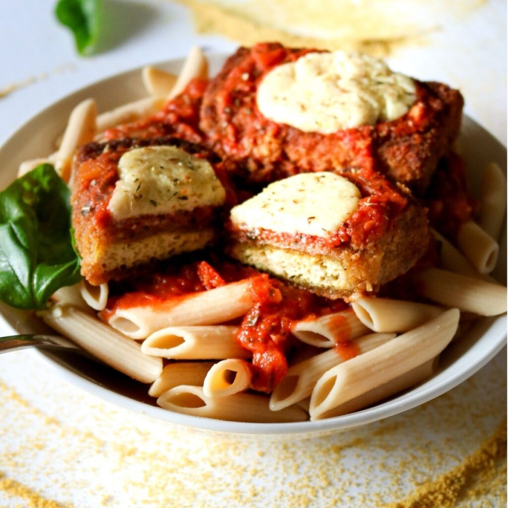 Overhead view of a round white bowl filled with penne pasta topped with saucy vegan chicken parmesan made out of tofu and sliced in half
