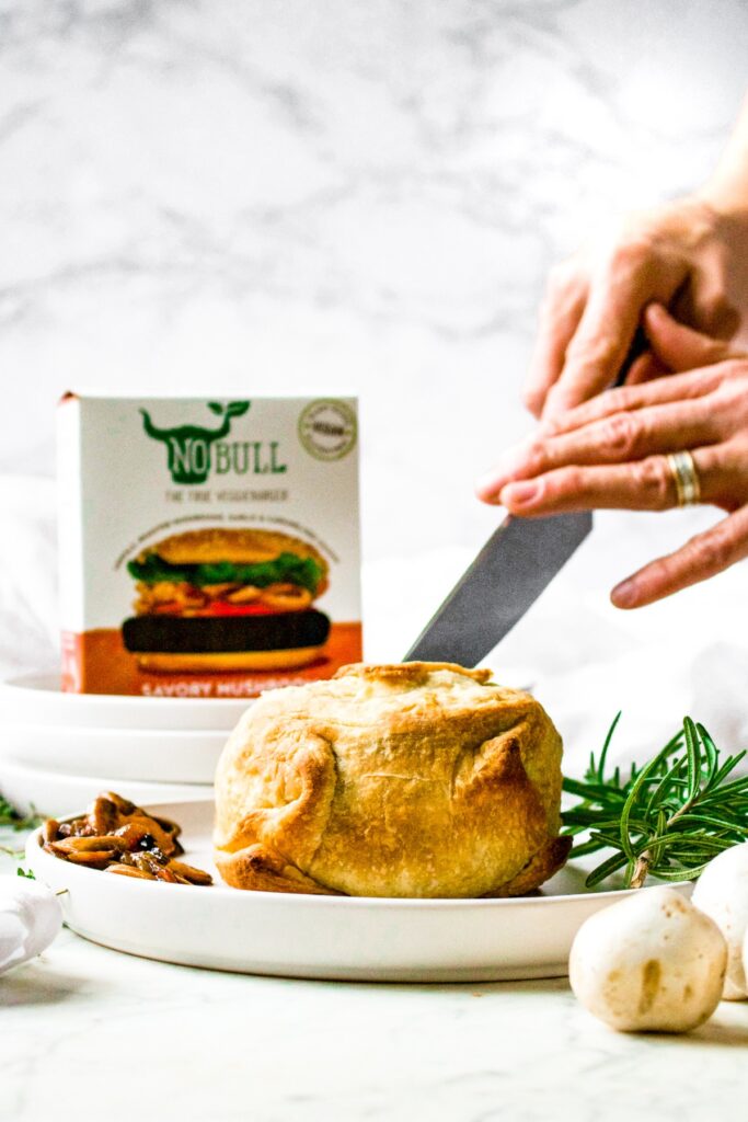 Head on shot of hands holding a knife and slicing into a Vegan Mushroom Wellington with mushrooms and a sprig of rosemary next to it