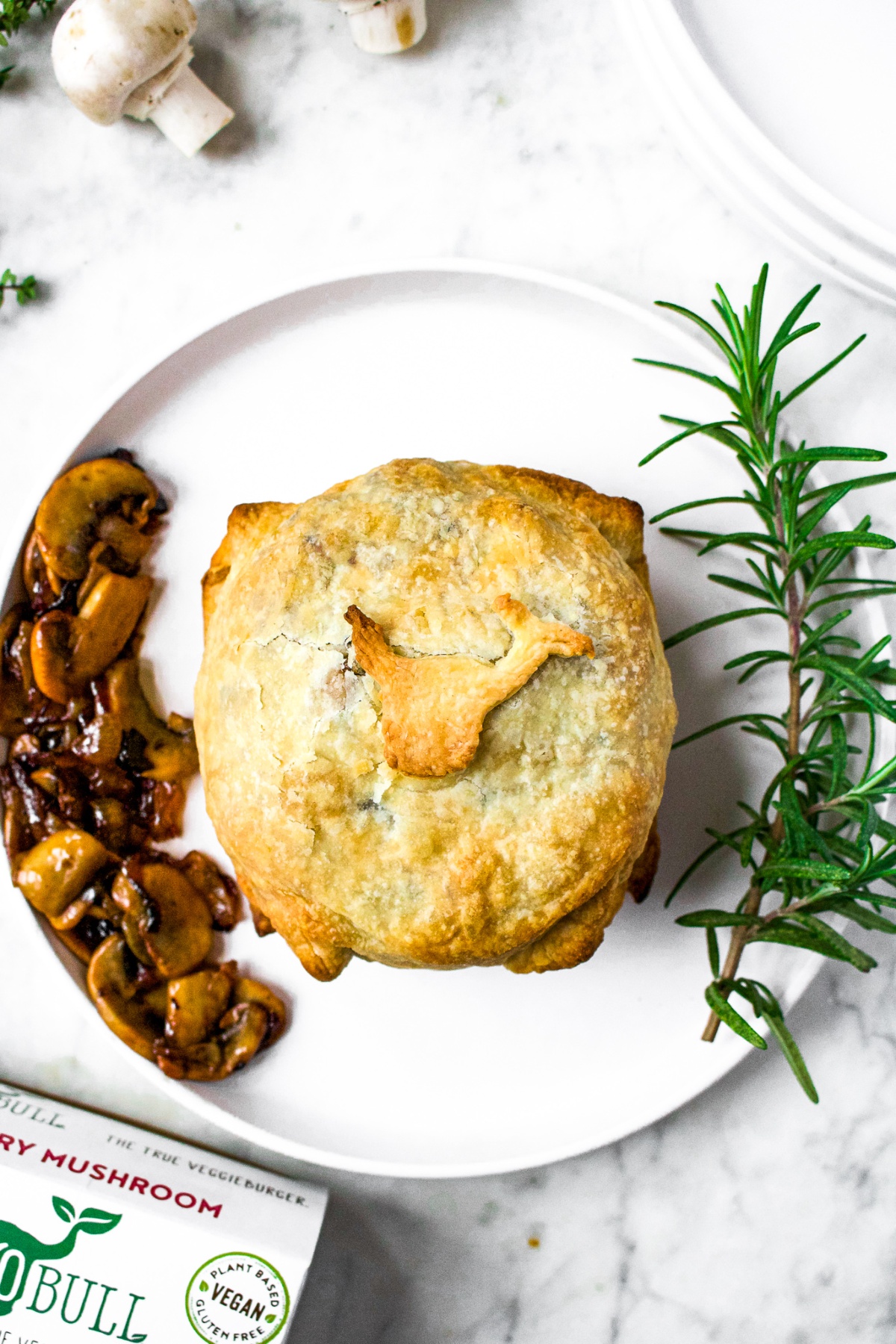 Overhead shot of a vegan mushroom wellington wrapped in crispy puff pastry sitting on a round white plate, surrounded by sauteed mushrooms and onions and a sprig of rosemary