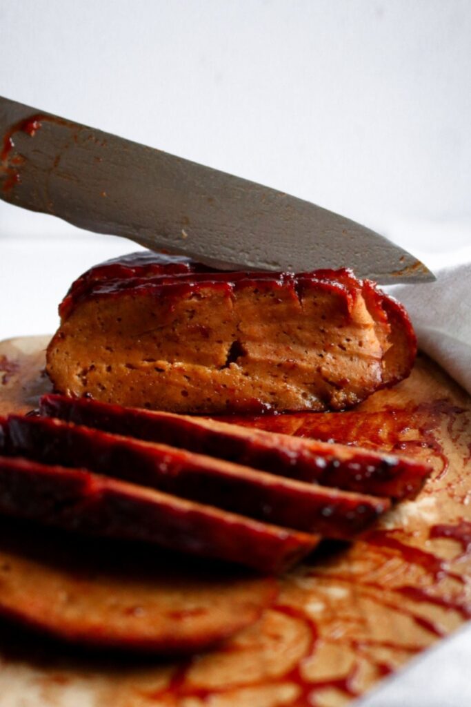 Head on view of a knife slicing into a vegan ham seitan roast on a wood cutting board