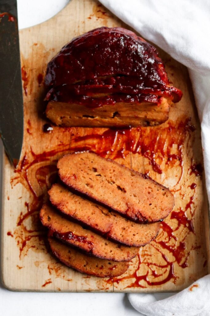 Overhead view of a vegan ham roast made of seitan half sliced on a wood cutting board surrounded by a thick messy pink-colored glaze