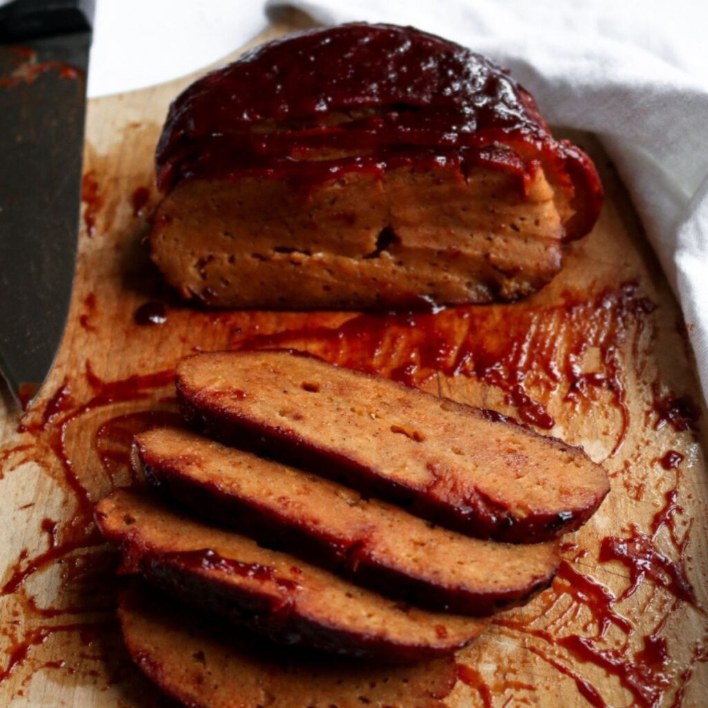 Overhead view of a partially sliced vegan ham roast on a wood cutting board with a thick pink pineapple glaze