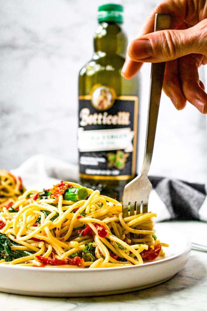 Head-on shot of a pile of spaghetti aglio e olio with vegetables on a plate with a fork digging in and twirling the pasta. A bottle of Botticelli Extra Virgin Olive Oil in the background.