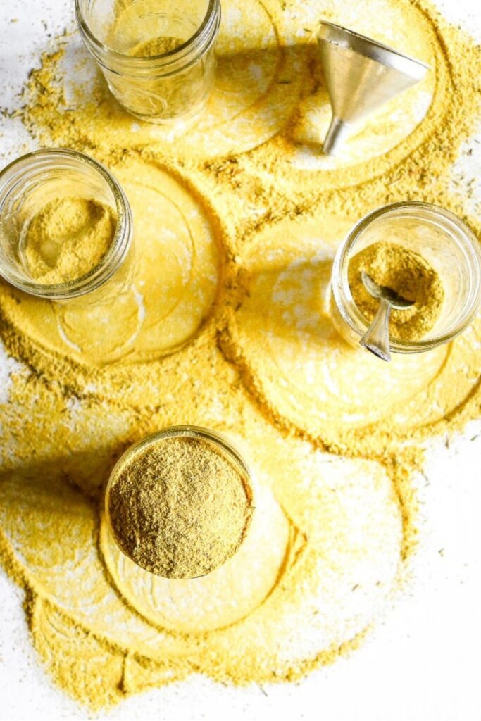 Overhead view of a white counter with vegan bouillon powder spilled everywhere in circular patters and three clear glass jars with a metal funnel