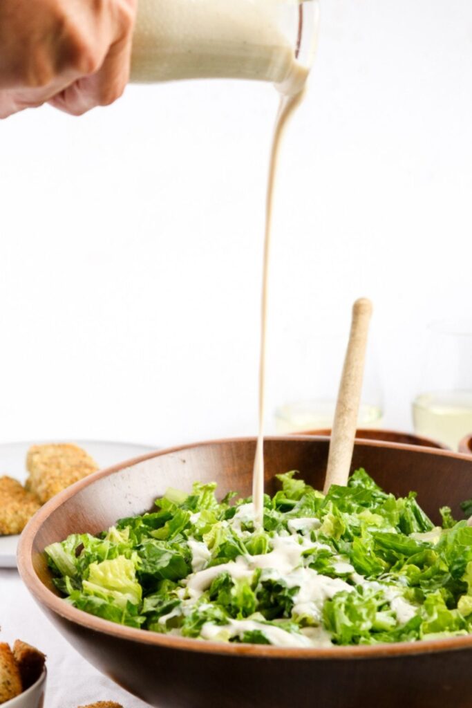 Head-on view of a hand pouring a glass jar of creamy vegan Caesar salad dressing onto romaine lettuce in a bamboo salad bowl