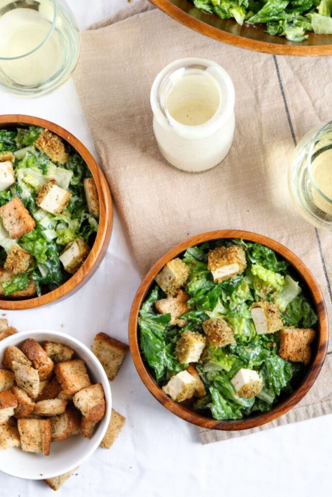 Overhead view of two round bamboo bowls filled with vegan Caesar salad topped with vegan lemon pepper tofu chicken next to a bowl of croutons and a glass jar of vegan caesar salad dressing