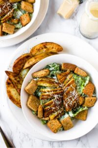 Overhead view of a round white bowl filled with a vegan chicken caesar salad topped with croutons and dairy-free parmesan next to a pile of potato wedges