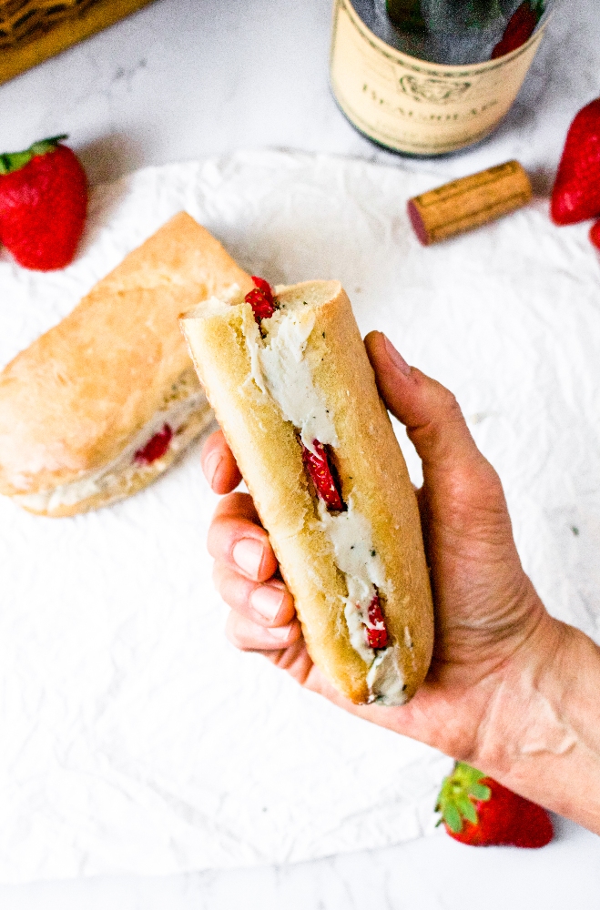 Overhead shot of a hand holding a Baguette sandwich with a vegan cashew cheese filling and sliced strawberries. There is another sandwich half, a strawberry, and wine cork in the background.