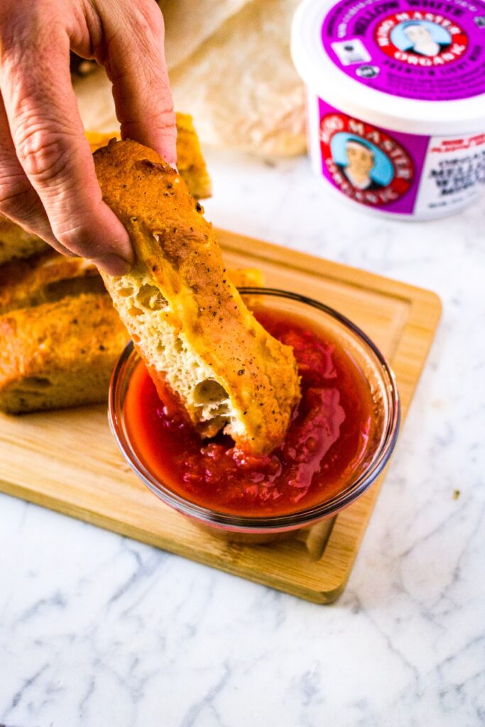 Close-up shot of a hand dipping a vegan parmesan breadstick into a small clear bowl of marinara sauce. There is a pile of cheesy breadsticks and a tub of Miso Master Mellow White Miso Paste in the background.