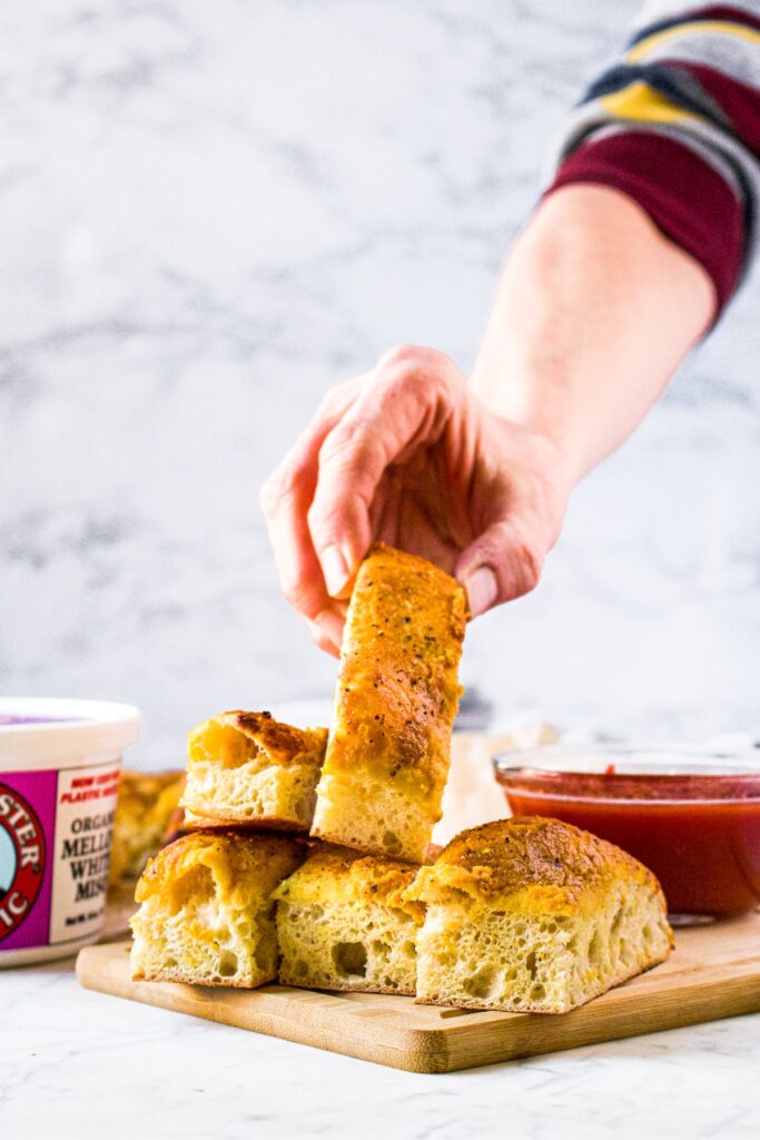 Head-on shot of a pile of vegan parmesan breadsticks. A hand is reaching in to pick up the top breadstick on the pile. There is a tub of Miso Master Organic Mellow White Miso Paste and a jar of marinara sauce in the background.