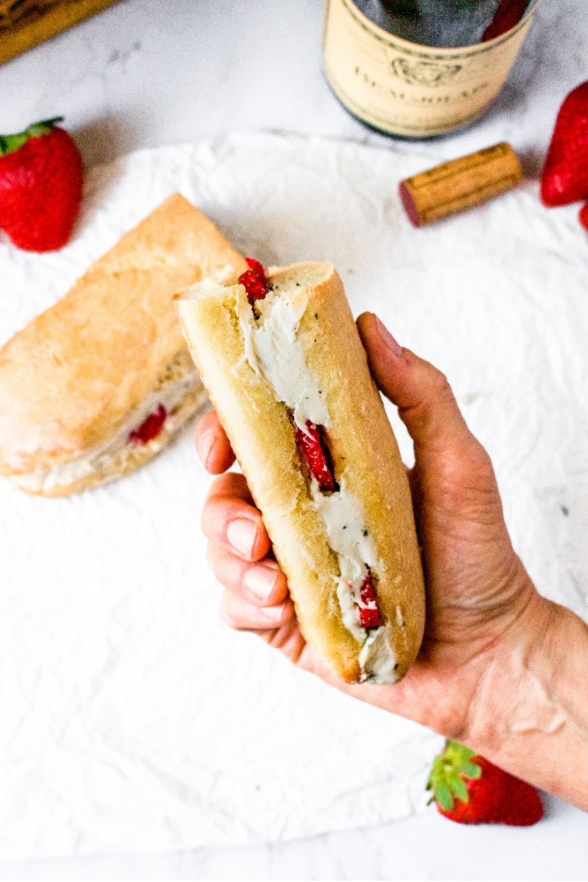 Overhead view of a hand holding a Baguette sandwich with a vegan cashew cheese filling and sliced strawberries. There is another sandwich half, a strawberry, and wine cork in the background.