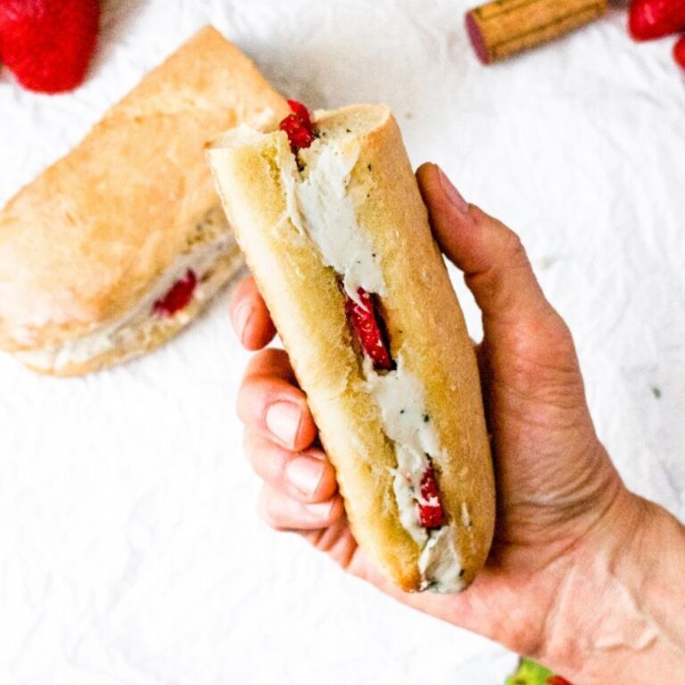 Overhead view of a hand holding a Baguette sandwich with a vegan cashew cheese filling and sliced strawberries. There is another sandwich half, a strawberry, and wine cork in the background.